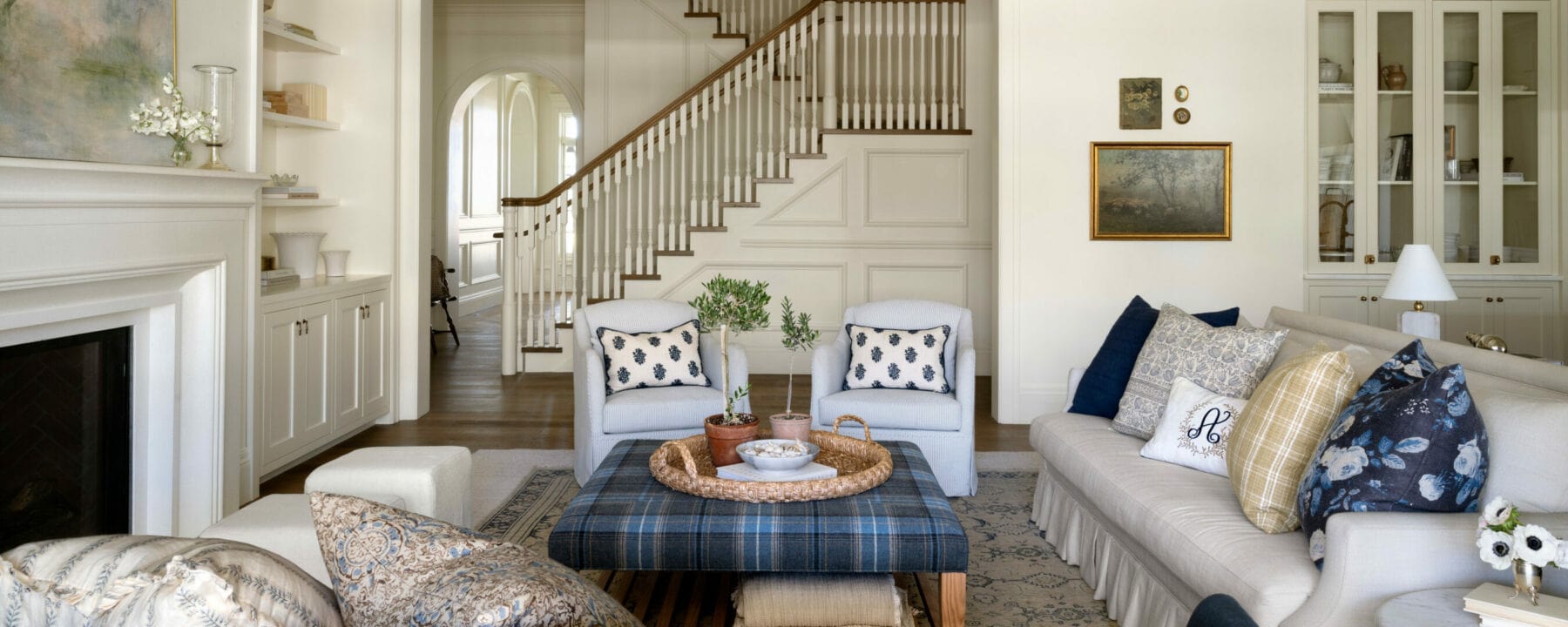 Elegant living room with neutral-toned sofas, blue plaid ottoman, decorative pillows, a wooden tray with plants, armchairs, framed art, and a staircase in the background, all in a light, airy setting, by Studio McGee.