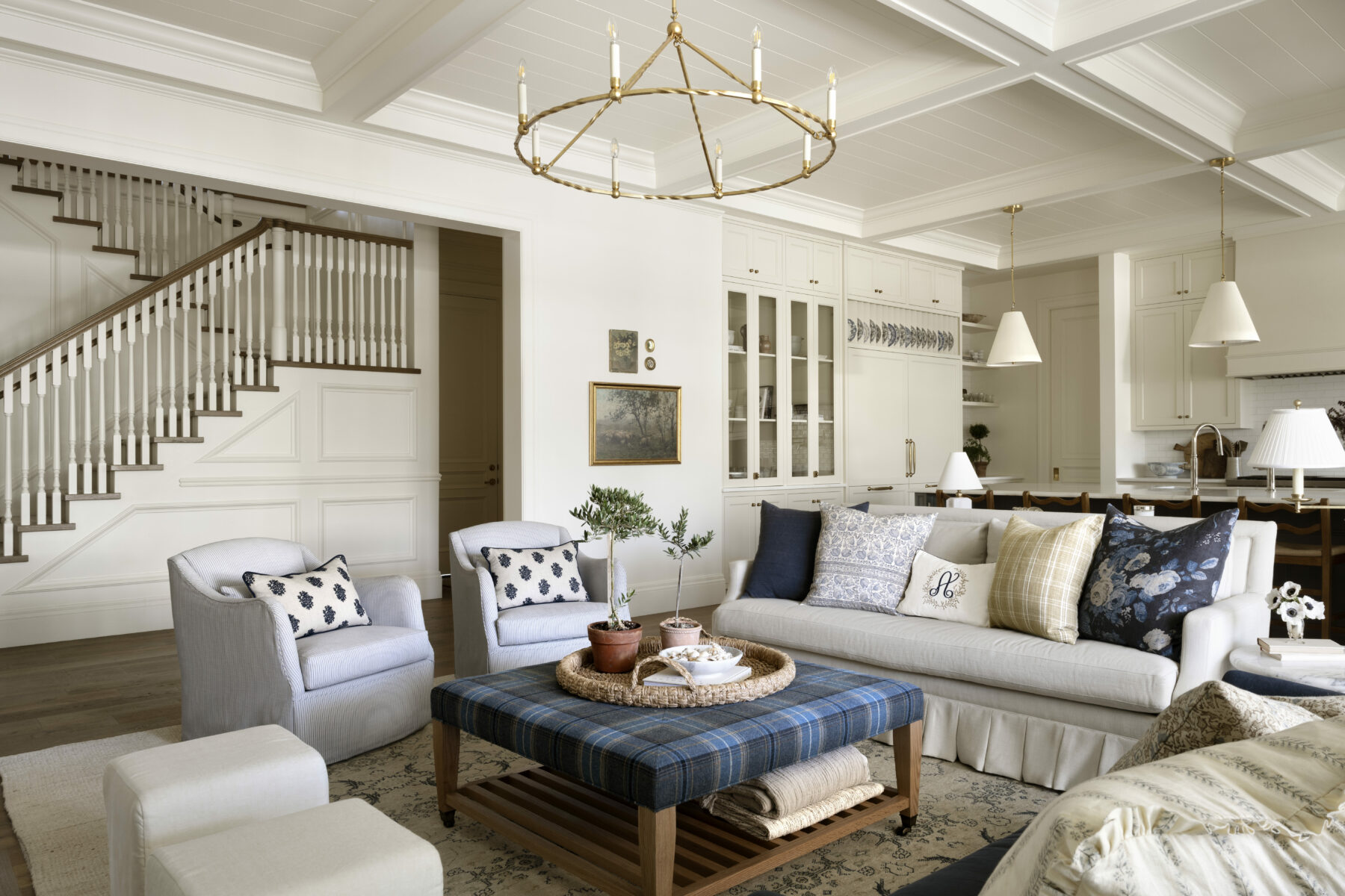 A bright, elegant living room with neutral walls, cozy armchairs, a white sofa with patterned pillows, a plaid ottoman as a coffee table, and a brass chandelier. A staircase and open kitchen are visible in the background, by McGee & Co.